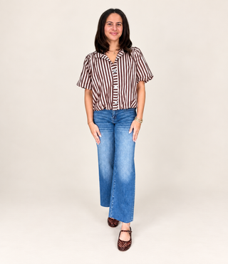 A woman modeling a brown and white vertically striped short-sleeve button-up shirt paired with blue wide-leg jeans and brown patterned flat shoes, standing against a plain light beige studio background.