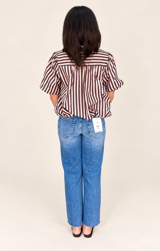 A woman viewed from behind wearing a brown and white vertically striped short-sleeve blouse tucked into blue jeans, standing against a plain beige studio background.