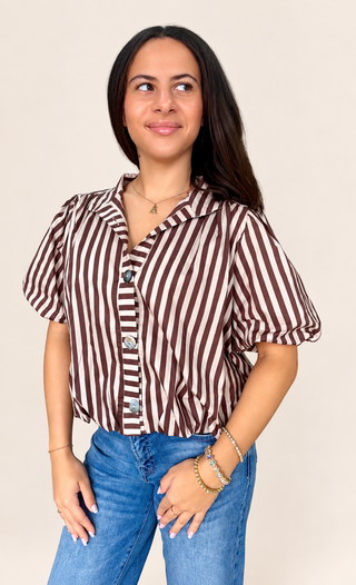 A woman wearing a brown and white vertically striped short-sleeve button-up shirt with puff sleeves, paired with blue jeans, standing against a solid light beige background.