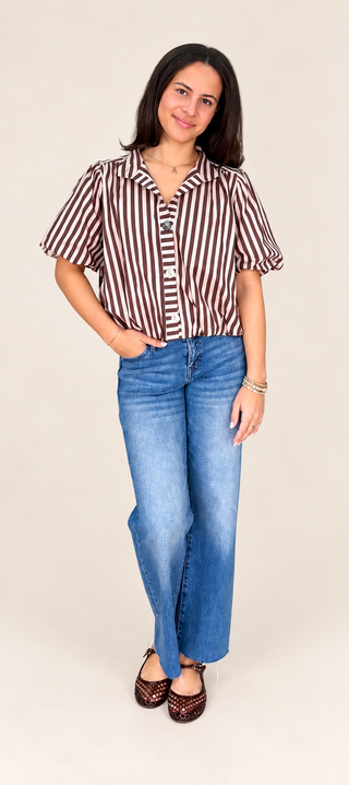 A woman modeling a short-sleeve button-up shirt with brown and white vertical stripes, paired with blue jeans and brown shoes, against a plain beige studio background.
