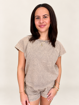 A woman modeling a matching two-piece lounge set consisting of a short-sleeve crewneck sweatshirt and shorts in a washed, mineral-washed taupe color, set against a plain light beige background.