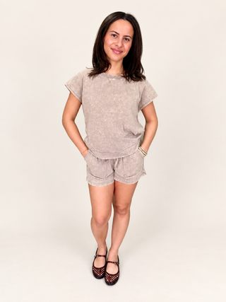 A woman modeling a matching two-piece linen set consisting of a short-sleeve t-shirt and shorts in a light beige color, paired with dark patterned flats, against a plain off-white studio background.