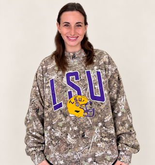 A woman wearing a camouflage crewneck sweatshirt with a purple LSU graphic and a yellow helmet logo on the front, set against a plain light beige background.