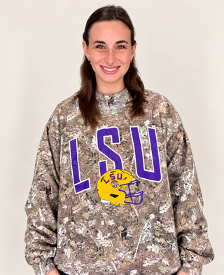 A woman wearing a camouflage long-sleeve sweatshirt with a purple and yellow LSU graphic on the front, photographed against a plain light beige background.