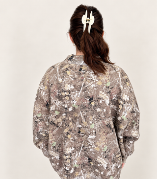 A person shown from behind wearing an oversized long-sleeve sweatshirt with an all-over camouflage-style floral and branch print in earthy beige, brown, gray, and muted green tones. The person has dark brown hair tied up with a white claw clip against a plain light beige studio background.