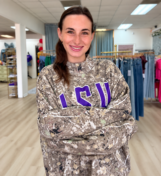 A woman smiling with arms crossed, wearing a camouflage-patterned sweatshirt with purple 'ICW' lettering on the chest, standing inside a retail store.