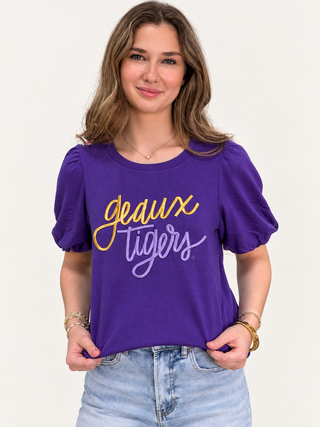 A woman wearing a purple short-sleeve t-shirt with 'geaux tigers' printed in yellow and purple script across the chest, paired with light blue jeans, against a plain off-white background.