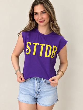 A woman wearing a purple sleeveless crop top with 'STTDB' printed in yellow across the chest, paired with light blue denim shorts, against a plain beige background.