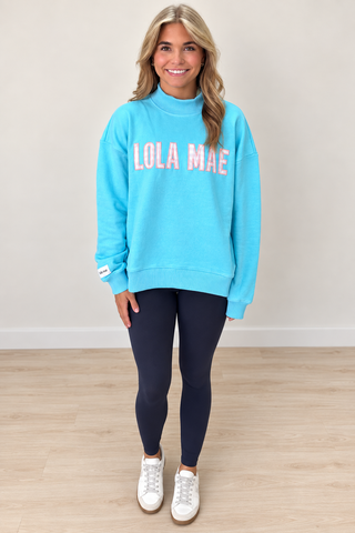 A woman modeling a bright turquoise blue mock neck sweatshirt with 'LOLA MAE' printed across the chest in pink and white, paired with dark navy leggings and white sneakers, standing against a neutral beige wall and light wood floor.