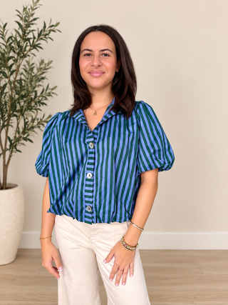 A woman modeling a blue and green vertically striped short-sleeve blouse with puff sleeves, paired with cream pants, standing in a brightly lit room with a potted plant and light wood floor.