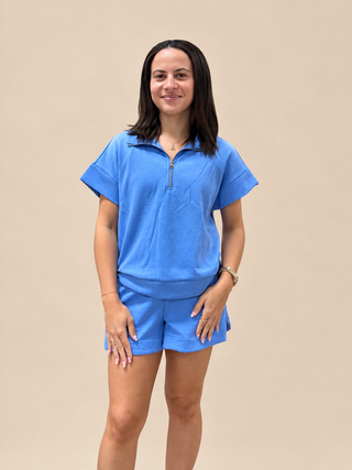 A woman modeling a matching blue lounge set consisting of a short-sleeve half-zip polo shirt and matching shorts, set against a solid beige background.