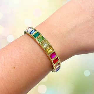 A gold-plated bracelet with rectangular, bezel-set rainbow gemstones worn on a person's wrist, set against a soft, blurred bokeh background.
