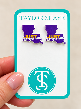 A hand holds a white display card featuring two purple Louisiana-shaped earrings with yellow and white 'JUST DIFFERENT' text. The card is branded 'TAYLOR SHAYE' with a teal circular 'TS' logo at the bottom.