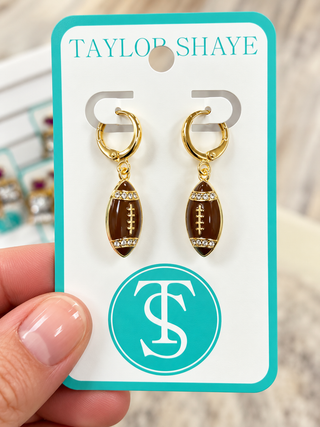 A pair of gold hoop earrings with football-shaped charms, held on a white display card with turquoise trim. A hand is visible holding the card, and the card is branded Taylor Shaye with a large TS monogram.