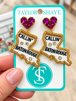 Taylor Shaye embroidered patch earrings shaped like the state of Louisiana, featuring a bright pink heart-shaped rhinestone stud and white, black, and gold embroidered details with 'CALLIN' BATON ROUGE' text, shown on a branded display card held by a hand.