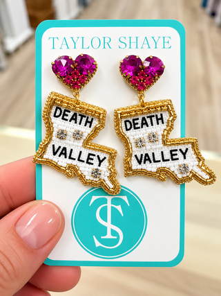 A hand holding a turquoise card displaying a pair of statement earrings shaped like the state of Louisiana, with 'DEATH VALLEY' text, gold-tone outlines, white textured centers, and bright pink heart-shaped rhinestones. The card reads 'TAYLOR SHAYE' at the top.