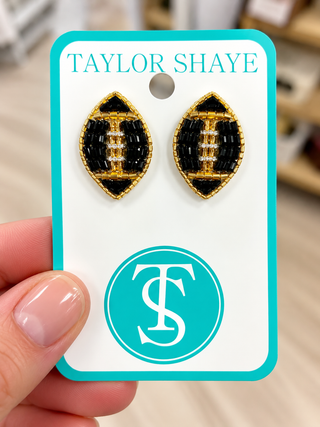 A pair of football-shaped stud earrings with black and gold beadwork, displayed on a white card with turquoise borders that reads 'TAYLOR SHAYE' and a large 'TS' logo. A hand is holding the card in front of a softly blurred indoor background.