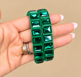 A hand with manicured nails holds a green rhinestone bracelet. The bracelet features a grid of square-cut green crystals set in a metallic green frame.