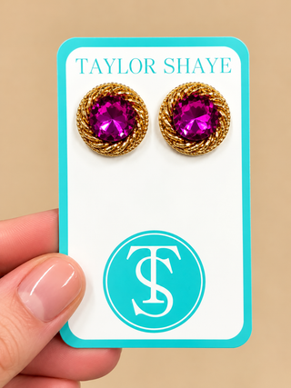 A close-up of a hand holding a jewelry display card featuring a pair of gold-toned twisted stud earrings with bright pink gemstones. The card is branded TAYLOR SHAYE with a teal logo and monogram.
