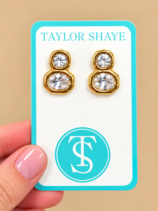 A close-up of a hand holding a jewelry display card featuring a pair of gold-toned stud earrings with clear faceted stones. The card is branded TAYLOR SHAYE with a turquoise logo and monogram, against a warm beige background.
