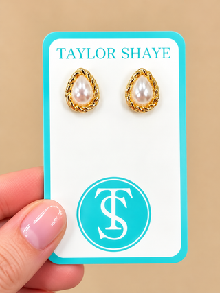 A pair of gold teardrop stud earrings with white pearl centers, displayed on a white card with a turquoise border. A hand with pink manicured nails holds the card against a solid beige background.