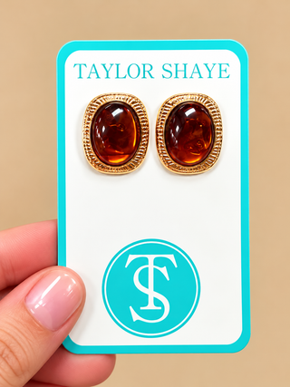 A pair of gold oval stud earrings with amber-brown cabochon centers, displayed on a white jewelry card with turquoise borders. A hand is holding the card against a beige background.