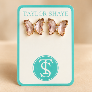 A pair of butterfly-shaped stud earrings with pink faceted crystals and gold-tone settings, displayed on a white card with a turquoise border. The card is branded 'TAYLOR SHAYE' at the top and features a matching circular 'TS' logo at the bottom, set against a neutral beige background.