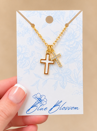 A gold cross necklace with a small crystal-embellished cross pendant, displayed on a decorative white card held by a hand. The card features a light blue floral pattern and the brand name 'Blue Blossom' printed at the bottom.