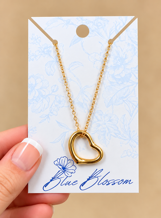 A gold open heart pendant necklace on a delicate gold chain, displayed on a white card with blue floral patterns and the brand name 'Blue Blossom'. A hand with a French manicure is holding the card on the left side, set against a solid beige background.