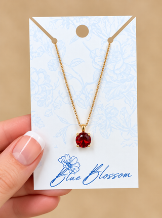 A gold chain necklace with a round red gemstone pendant, displayed on a white card with blue floral details and the brand name 'Blue Blossom'. A hand with a French manicure is holding the card against a solid beige background.