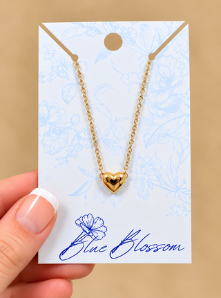 A gold heart pendant necklace on a delicate gold chain, displayed on a white card with blue floral patterns and the brand name 'Blue Blossom'. A hand with a French manicure is holding the card on the left side, set against a solid beige background.