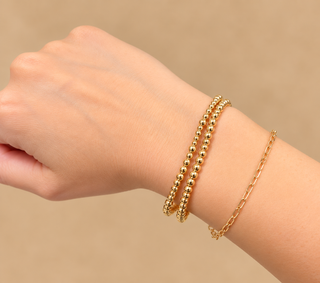 A close-up of a person's wrist wearing three gold bracelets: two beaded bangle bracelets and one delicate chain bracelet, against a soft beige background.
