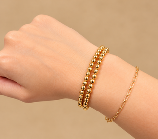 A close-up of a person's wrist wearing two gold bracelets: one with small metallic beads and one with a delicate chain link design, against a soft beige background.