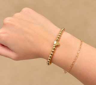 A close-up of a person's wrist wearing two delicate gold bracelets against a soft beige background. One bracelet features a small polished gold heart charm, while the other is a fine gold chain.