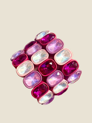 A wide cuff bracelet featuring multiple rows of iridescent and deep pink cabochon gemstones set in a metallic pink base, shown against a solid light beige background.