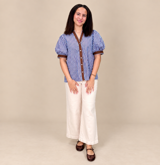 A woman modeling a blue and white striped short-sleeve blouse with brown trim, paired with off-white wide-leg pants and dark brown shoes, against a solid beige studio background.