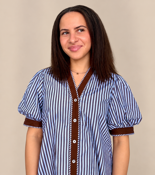 A woman with shoulder-length dark brown hair wearing a blue and white vertically striped short-sleeve blouse with brown trim and white buttons, photographed against a plain beige studio background.