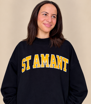 A woman wearing a black crewneck sweatshirt with 'ST AMANT' printed in yellow varsity-style letters with a white outline across the chest, set against a plain beige background.
