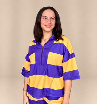A woman wearing a purple and yellow striped athletic half-zip top, standing against a solid beige studio background.