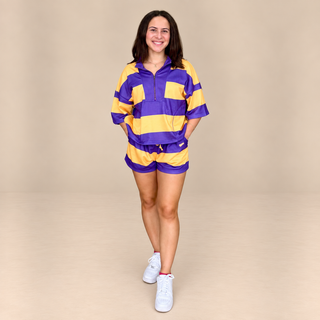 A woman modeling a matching purple and yellow striped athletic set consisting of a half-zip pullover and shorts, paired with white sneakers, against a solid beige studio background.