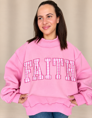 A woman wearing a pink mock-neck sweatshirt with the word 'FAITH' in pink-and-white gingham block letters outlined in black, posed against a neutral beige background.