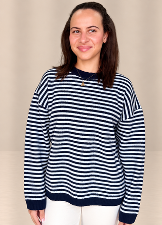A woman wearing a navy blue and white horizontally striped knit sweater, paired with white pants, standing against a plain beige studio background.