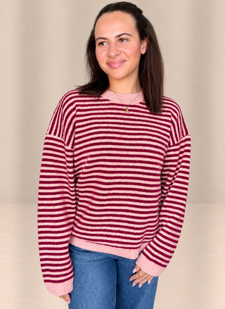 A woman wearing a fuzzy long-sleeve sweater with horizontal burgundy and pink stripes, paired with blue jeans, standing against a neutral beige background.