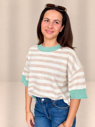 A woman wearing a beige and white horizontally striped knit top with mint green ribbed collar, cuffs, and hem, paired with blue jeans and sunglasses on her head, set against a plain beige background.