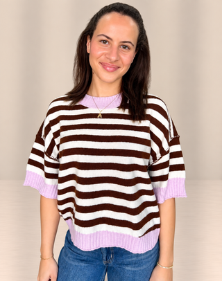 A woman wearing a brown and white horizontally striped knit sweater with light purple ribbed trim at the crew neck, sleeves, and hem, paired with blue jeans.