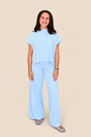 A woman modeling a light blue two-piece lounge set consisting of a short-sleeve mock-neck top and matching wide-leg pants, standing against a solid beige background.