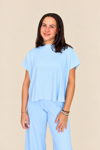 A woman modeling a light blue short-sleeve crop top and matching light blue pants against a solid beige studio background.