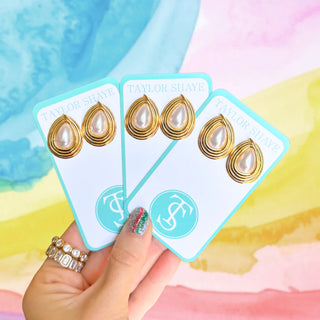 Three pairs of gold and pearl teardrop earrings on Taylor Shave cards held by a hand against a colorful background.