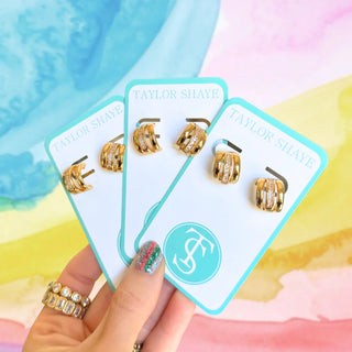 Gold earrings on Taylor Shaye cards held by a hand with colorful nail polish against a multicolored background.