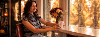 A woman in a striped top sits by a window in a warmly lit indoor setting, holding a white mug next to a vase of flowers.